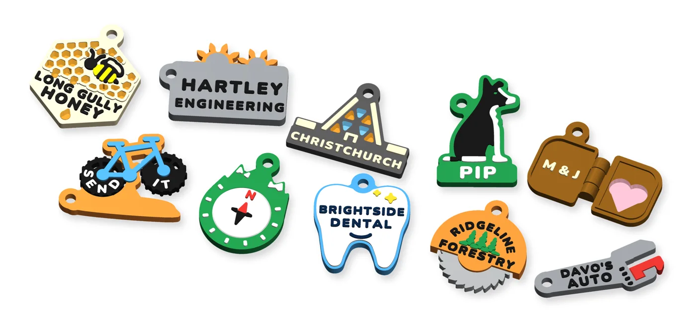 Ten one-off keychains scattered on a white table, each in its own colours: a honeycomb with translucent honey, two gears, a stained-glass cathedral, a border collie, a mountain bike, a compass dial, a glossy tooth, a hinged locket with a heart window, a saw blade and an adjustable spanner.